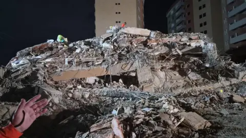 UK International Search and Rescue A collapsed structure with a hand pointing towards it in the bottom left. There is lots of rubble and behind are tower blocks. 