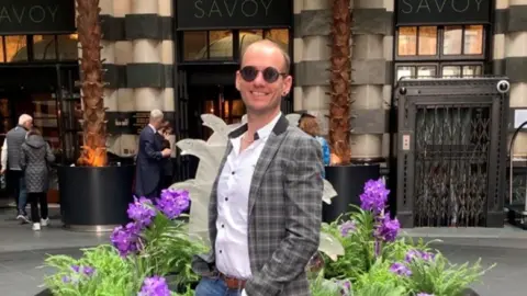 Ben Howard Ben Howard is pictured out in the street in front of a flower bed. He is wearing a pair of round sunglasses and grey checkered jacket over a white collared shirt. He is smiling at the camera. 