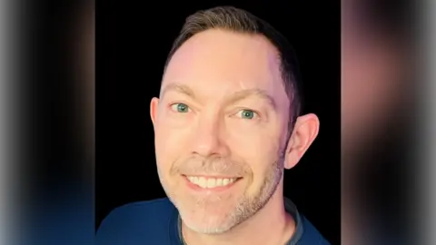 A man with short black hair and a blue top smiles as he stands in front of a black background.