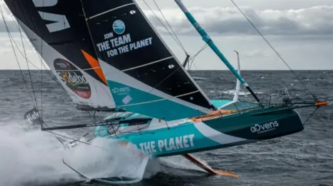 Pierre Bouras A racing yacht with a white and blue hull and black and white sails going fast with spray behind.