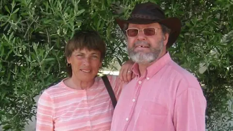 Family photograph Mary and Noël Hansen, with a bush behind them, posing for a holiday photograph. She has short brown hair; he is wearing a small black stetson hat.