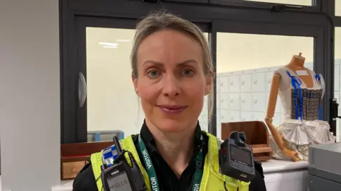 A police officer with blonde hair and blue eyes is stood in front of a window in a classroom. She has a florescent yellow jacket on with two radio attached. A mannequin with no head is in the background, wearing a white dress.