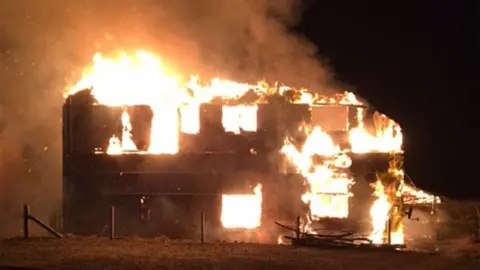 A building completely engulfed by fire. There is no roof, flames and smoke are billowing out of windows, against a black sky.