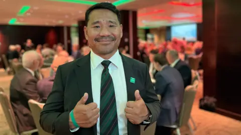 Hari smiles at the camera. He is wearing a jacket with a white shirt and a tie with dark green and black diagonal strips. He has a green wristband on his left hand and a Gurkha Welfare Trust badge on his jacket. Behind him people are sitting at circular tables eating dinner in a large dining room.  