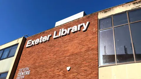 A white sign for Exeter Library on a red brick wall with windows to either side.