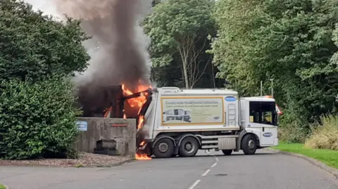 A Durham County Council bin lorry on fire. Flames engulf the rear of the lorry, which has turned parked across two lanes of a road.