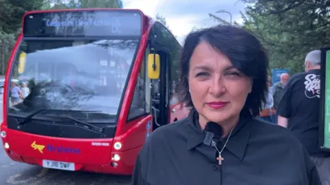 A woman with black hair, wearing a black shirt, standing next to a red bus at a bus stop.