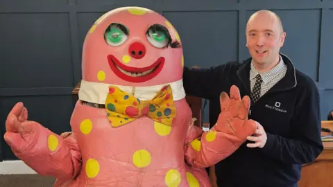 Auctioneum Ltd A man wearing a fleece jacket is standing next to a large, pink costume is pictured with big googly eyes, a yellow bow tie and dots.
