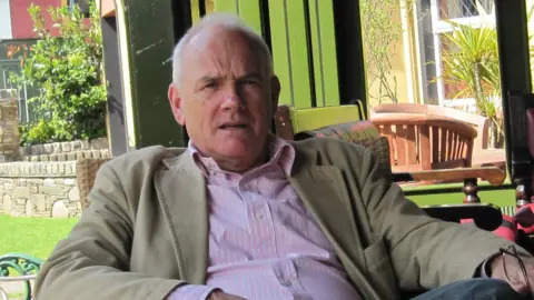 a man in a shirt and blazer reclining slightly in a wicker chair, with a garden visible in the background