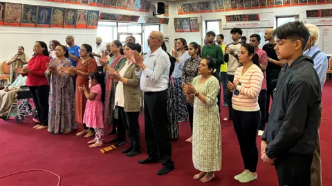 A group of about 30 people stand in a large red-carpeted room. A boy in the foreground has his eyes closed and hands clasped in front of him. Other people are singing and clapping their hands.