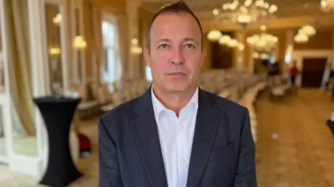 A man facing the camera wearing a white shirt with a blazer over the top. Background of a hotel meeting room is blurred.