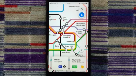 A mobile phone on a Tube carriage seat displaying a close up of part of the Tube map on the app, with status disruption and payments history options visible at the bottom of the app. 