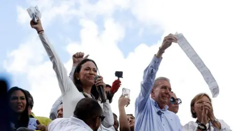 Opposition candidate Edmundo González, wearing a blue shirt, and opposition leader María Corina Machado, wearing her trademark white T-shirt, both hold aloft voting tallies from the presidential election. They are surrounded by several people who clap and cheer them on.