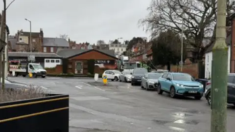 Claire Ellis A line of cars on a road with houses and buildings nearby.