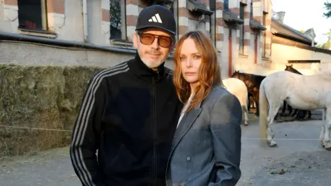 Alasdhair Willis is wearing a dark blue cap and tracksuit top and a pair of sunglasses with brown lenses. Stella McCartney has shoulder length brown hair and is wearing a grey suit jacket. The couple are standing outside a large building and there are four horses in the background.