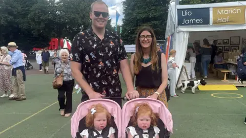 James Cook has blonde short hair, wearing sunglasses and a black patterned short-sleeved shirt. He is tall and standing next to Nerys Cook who  has long brown hair and is wearing a turquoise chunky necklace, a black tank top and an orange patterned skirt. In front of them is a pink double pram with ginger-haired twins Lili-Wen and Gwenllian who both have a white bow on the left side of their head