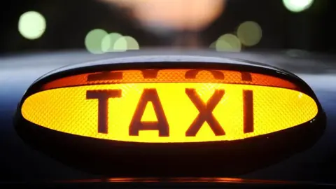 A lit, yellow taxi light on the top of a black cab. The word 'TAXI' is in black, and the rest of the light is yellow. Blurred out street lights are visible in the background.