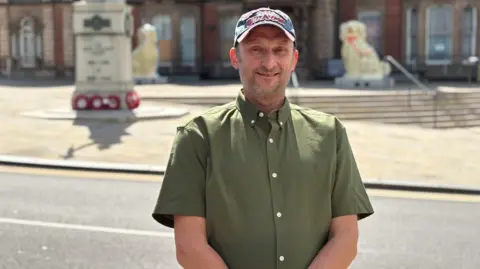 A man with a blue denim hat on is smiling and wearing a khaki green shirt. He is standing in front of a road with a war memorial on the adjacent pavement with poppy wreaths at its base