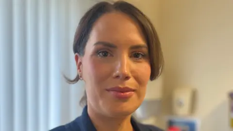 Dr Jilly O'Hagan has brown hair parted in the middle and tied back in a pony-tail and has brown eyes. She is wearing a navy suit jacket