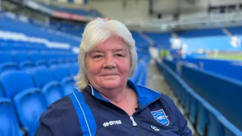 A woman with short white hair is wearing an old-style blue Brighton & Hove Albion FC jacket. She is sitting in an empty football stand in a stadium; blue seats are in the background.