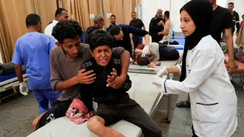 A boy sitting on a bed crises, while a man sat beside him hugs him and a woman stands beside him