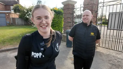 A young woman with blonde hair and glasses stands near a middle-aged bald man wearing black clothes and a gate
