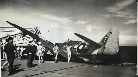 Joseph Douglas Cheshire A plane slanted on its side next to a crane on the landing platform of an aircraft carrier out at sea. Men are inspecting some with no tops on in the sunny weather