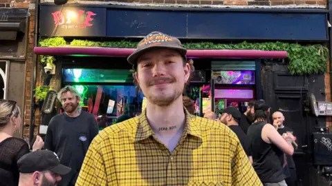 A man wearing a yellow checked shirt and baseball cap stands outside a music venue called Dive, with several other people gathered near the entrance.