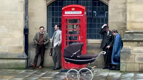 Five people dressed in period clothing standing next to a red phone box with a traditional pram in front of it 