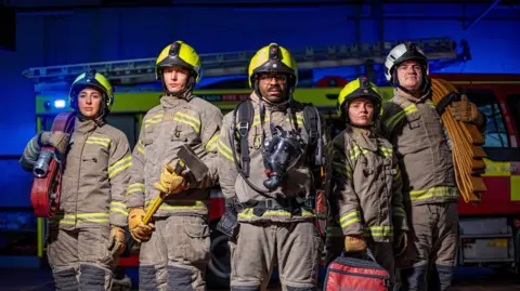 West Midlands Fire Service Five firefighters in brown fire suits with green reflective strips are standing in a line, in front of a fire engine. They are all wearing helmets. Each are holding a piece of kit, including hose reels and an axe.