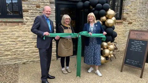 Three people holding a green ribbon on gravel floor outside art studio, with black and gold balloons in the background