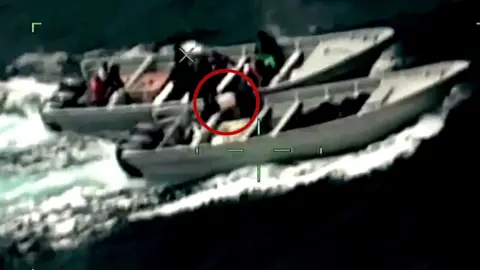 Two figures in black carry a large white bag circled in red between two motorboats from the point of view of a camera scope.