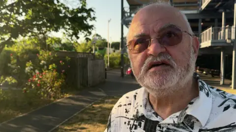 Dr Wael Elgonaid gives a TV interview in the street. He is bald with a white beard and wears a patterned black and white collared shirt and sunglasses.