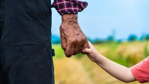 A photograph of an older man's hand holding the hand of a child with heavily blurred long green grass in the background.