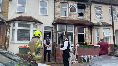  In the image, a firefighter and two police officers stand outside a terraced house on Napier Road in East Ham, where the ground and first floors show signs of a recent fire. The house has broken windows and a partially charred exterior. A man in a maroon shirt and other people are seen nearby, and there are flowers placed against the front wall as a tribute.