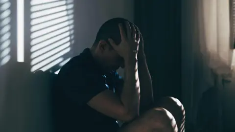 A stock image of a man holding his head in his hands crouched down against a wall. The image is quite dark in colour.