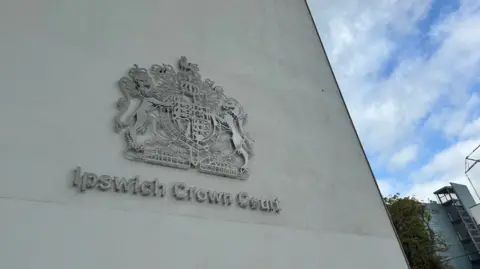 George King/BBC The royal coat of arms is fixed to a modern concrete building, with the words Ipswich Crown Court underneath. The camera is facing upwards at the crest, from the ground looking up. There is a blue sky and some white clouds visible to the right of the frame.