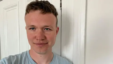 Éanna Mac Cana is smiling at the camera while standing in front of a white door and white wall. He has short, light brown hair and is wearing a light blue t-shirt.