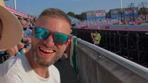 Simon Felton A man with a beard and brown hair is wearing sunglasses and on the left of the image smiling at the camera. He is at an athletics stadium and other people are sitting in seats in the background.