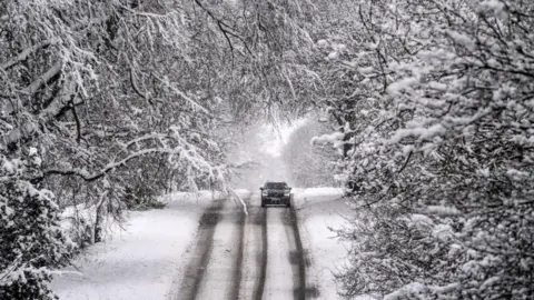 PA Media A car drives along an icy road, with snow-covered trees on either side.