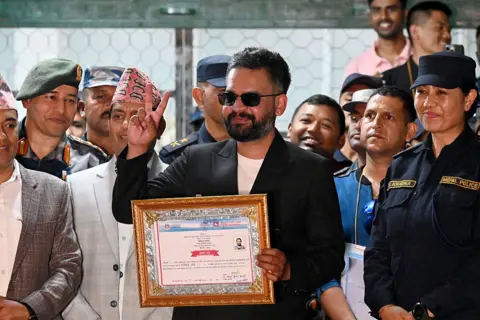 Balen Shah makes a peace sign with his fingers, and smiles to the crowd, while holding up a certificate 