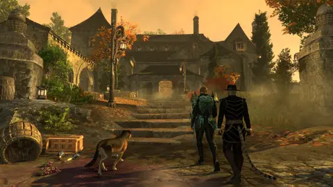 Bethesda Screenshot from Elder Scrolls Online shows two humanoid characters - one human and one with a tail - approaching a large, manor-house style building at sunset. Some fog lines the approach, which is littered with barrels and similar items, and a big cat companion walks alongside them.