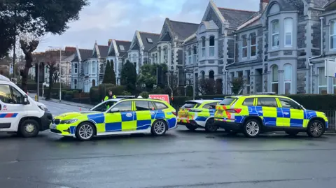 Police cars are seen blocking a road in Plymouth. There are a row of terrace houses behind them.
