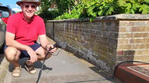 Guy Hargreaves has sunglasses on, as well as a red polo shirt and blue shorts. He is holding an electric car charger, with the lead running through a small, narrow trench in the path.