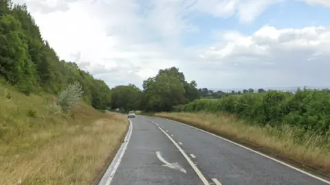 Google maps street view image of the A48 in Gloucestershire. One car can be seen in the distance on the road. There are hedges and trees either side of the carriageway. 