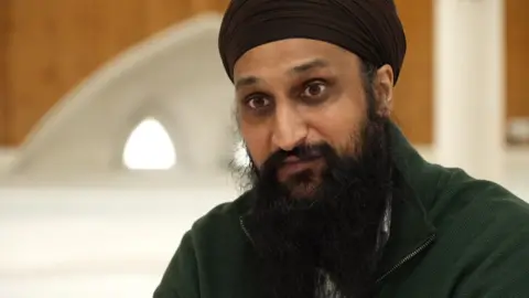 Harjit Singh, a man with a black beard wearing a black fleece and turban, is pictured facing the camera.