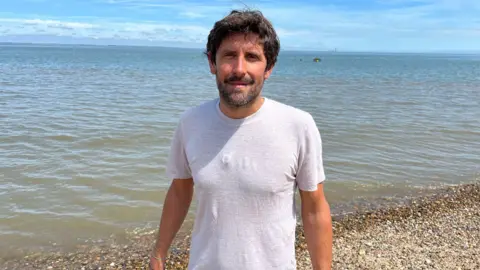 PHIL HARRISON/BBC A young man with dark hair and a beard wears a white t-shirt as he stands on a shingle beach close to the water.