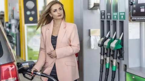 A woman with long blonde hair and wearing a pale peach-coloured jacket filling up her car at a petrol station 