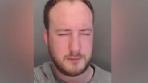 Mugshot of Henderson. He is 30 with light brown hair, a brown beard and is wearing a grey jumper. His eyes are closed.