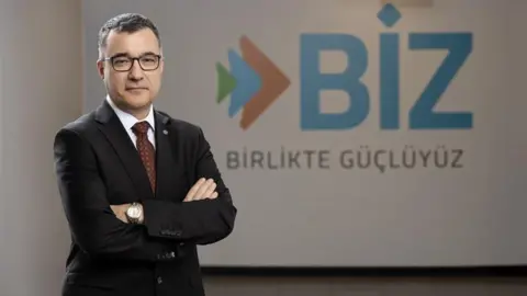 Contributed Ihsan Tolga Akar, wearing a dark suit, stands in front of a logo which says BIZ. He is wearing glasses, has his arms folded and has short grey hair. 